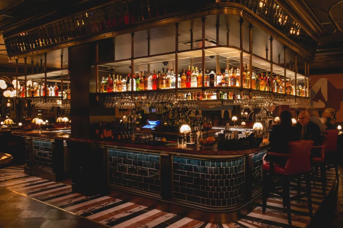 A picture of a steakhouse bar with blue tiling, warm tones, and an assortment of alcoholic bottles lit up on the shelves hanging above. 