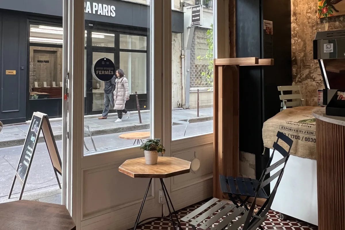 Inside Cafe Le Marais in Paris, looking out through the windows and open door at the street, with a small, wooden table and chair inside. 