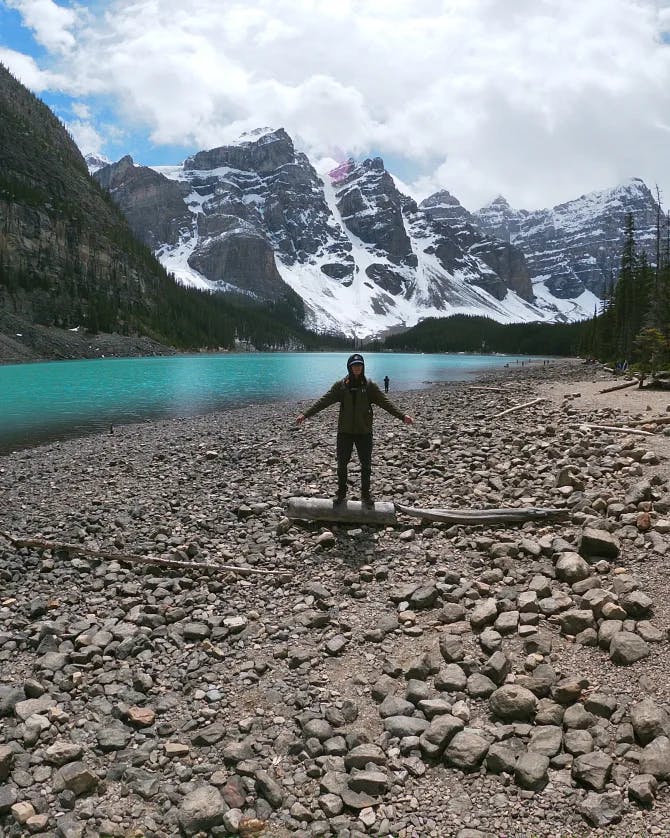 A picture of Tommy standing on rocks with a turquoise lake and snow covered mountains in the background