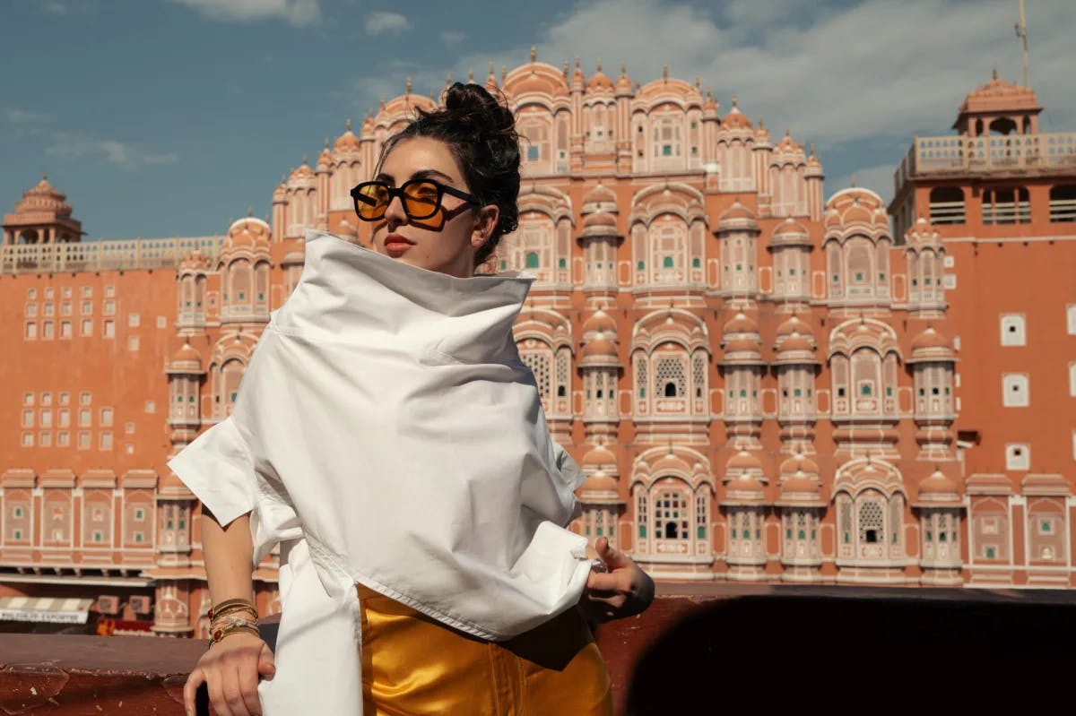 A woman posing in front of a luxurious pink building large in size, while wearing a silky gold skirt, sunglasses and white top. 