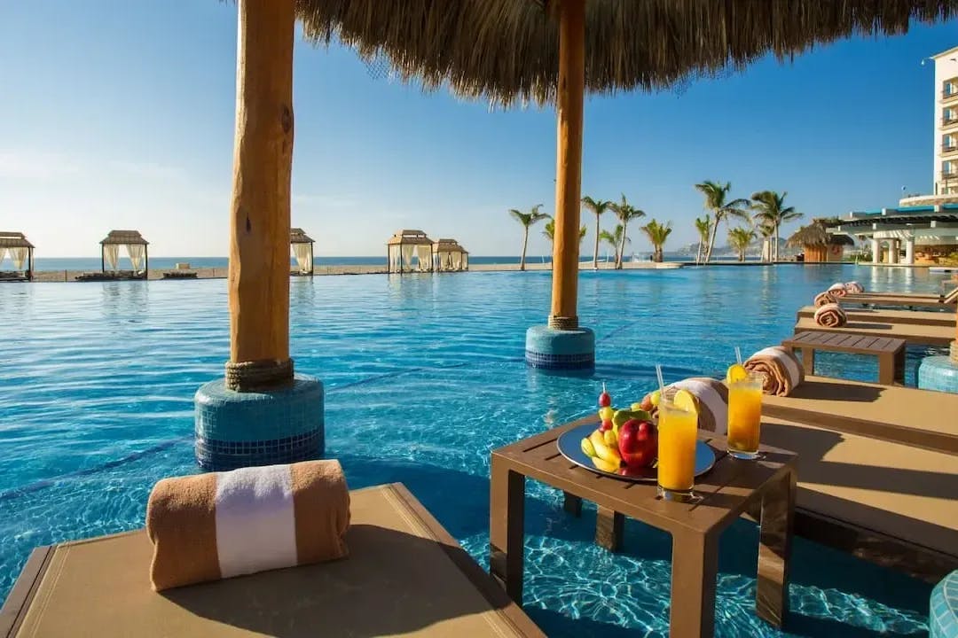 Loungers sit directly in the water of an infinity pool at Hyatt Ziva Los Cabos, with tropical cocktails and fruit on a serving dish next to one of the chairs