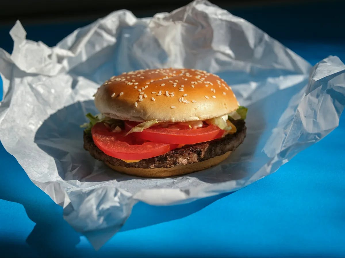 The image depicts a hamburger with lettuce and tomato on a sesame seed bun, wrapped in white paper on a blue surface.