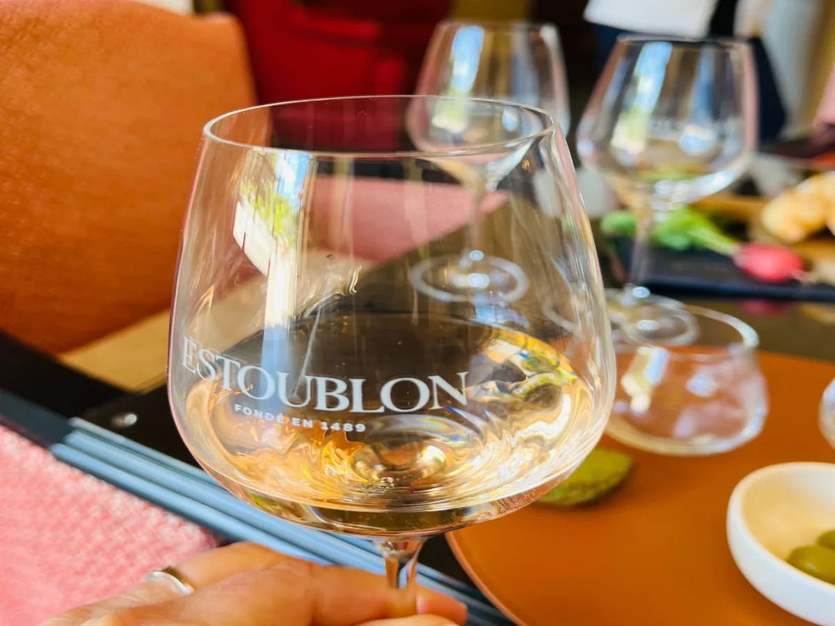 A close-up of a wine glass etched with "Estoublon,” with other glasses and a blurred social setting in the background.