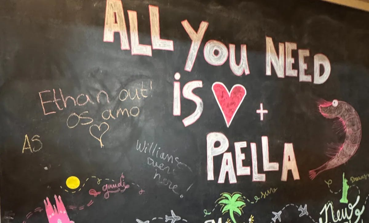 A chalkboard with the phrase “ALL YOU NEED IS LOVE + PAELLA” in colorful chalk, surrounded by playful doodles and names.