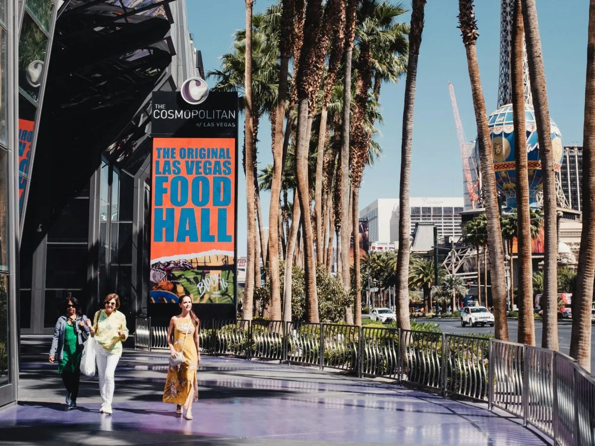 A group of people walking down a sidewalk next to palm trees with a sign behind them that reads "The Original Las Vegas Food Hall"