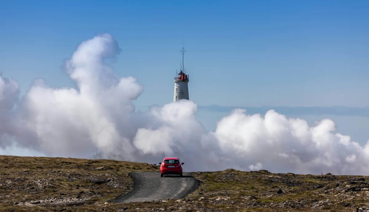 A red car driving toward the Gunnuhver spring, with a lighthouse and clouds.