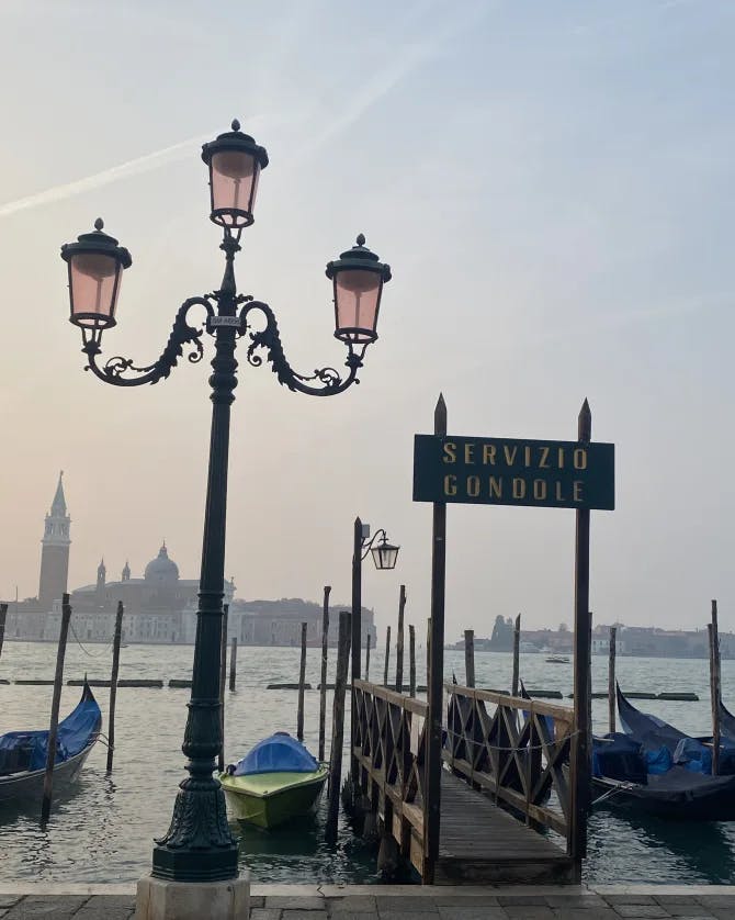 A boat dock in Venice with a sign that says "Servizio Gondole" with the Venice skyline in the background across the water.