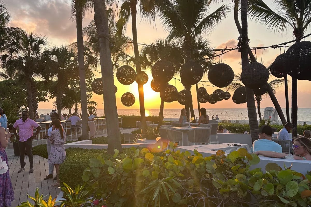 People standing around on a patio, with lanterns strung between palm trees and the ocean and sunset in the distance.