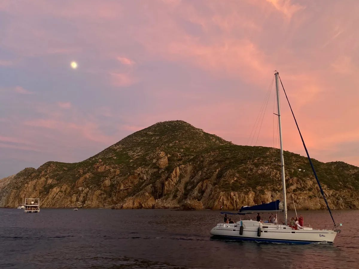 A view of a white sailboat in front of a rocky mountain above the water with the moon and a pink sunset in the background. 