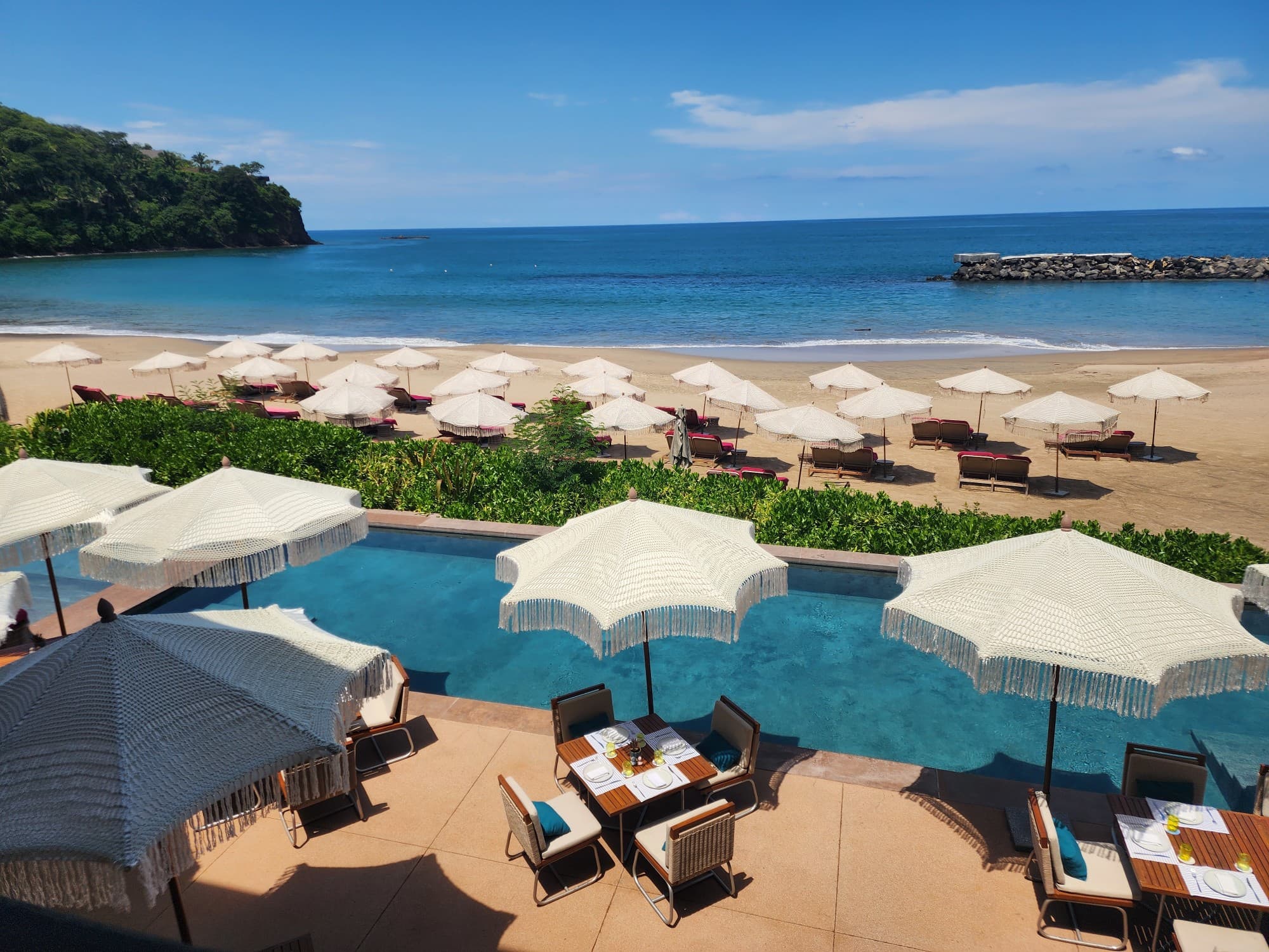 A view overlooking the pool with tables and chairs with umbrellas surrounding it and the beach with loungers and a calm ocean in the distance on a sunny day.