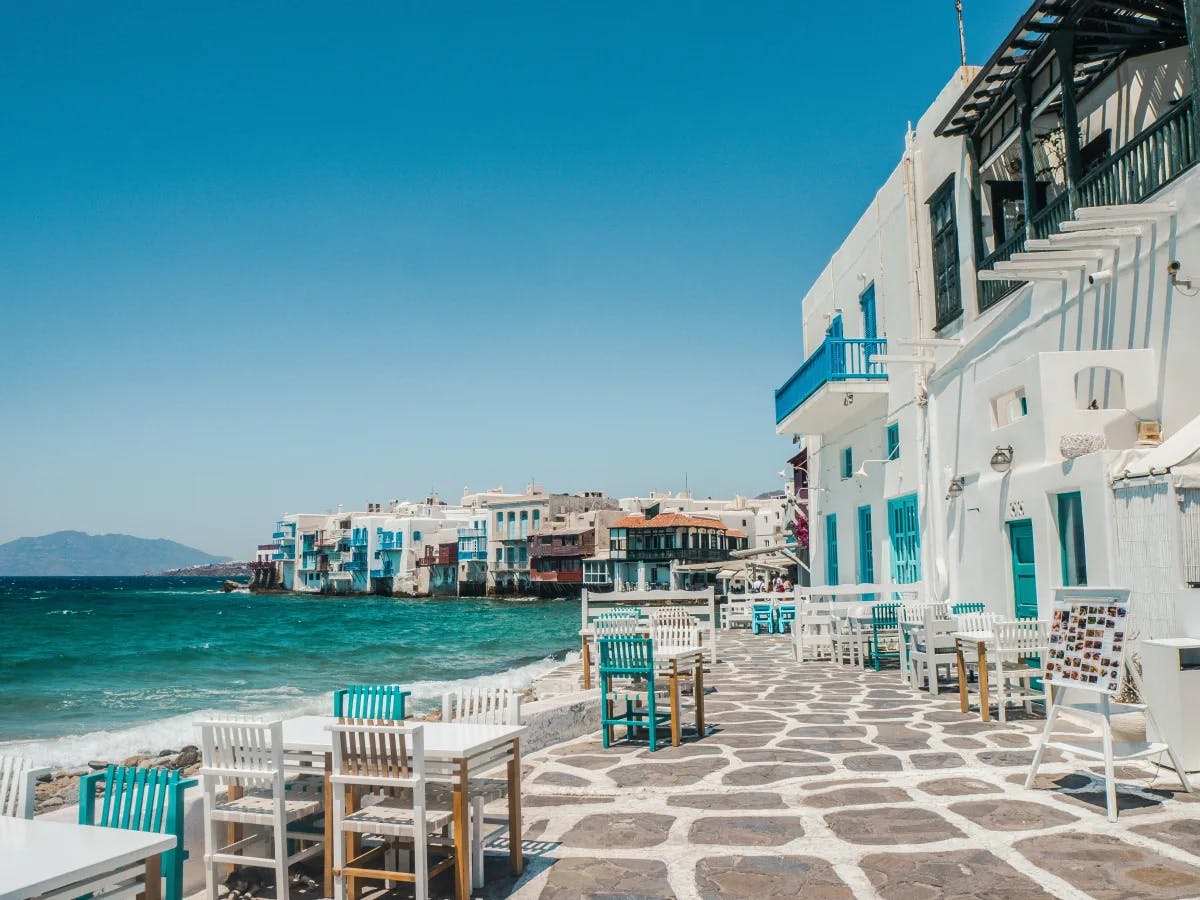 An outdoor patio with stone tiling, white blue and brown chairs and tables, and turquoise blue water with buildings in the surrounding areas. 