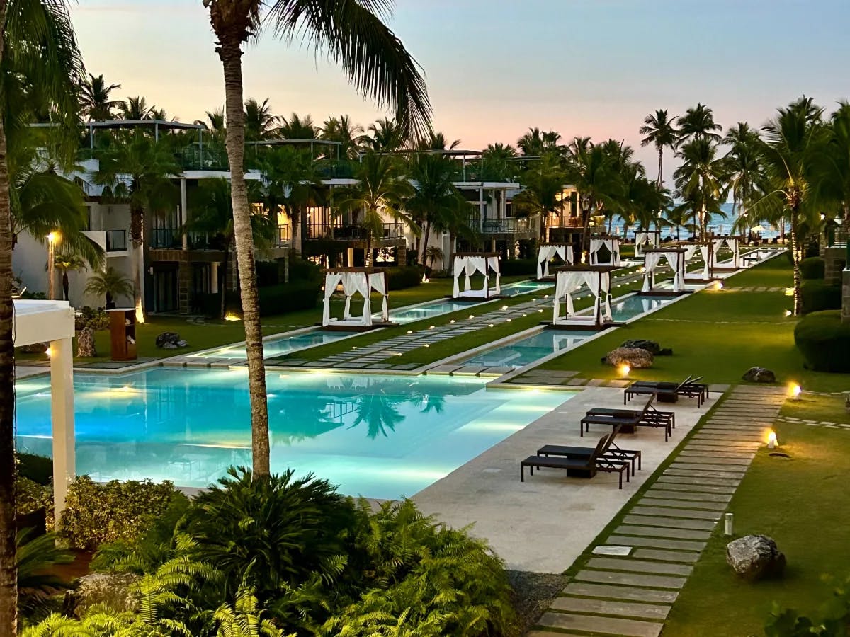 A view of the pool deck complete with decorative lighting, grass, palm trees, lawn chairs and white cabanas at sunset. 