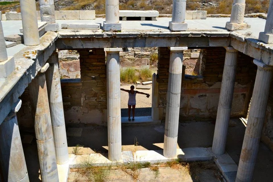 A girl standing with her arms outstretched in between pillars from an ancient Greek ruin. 