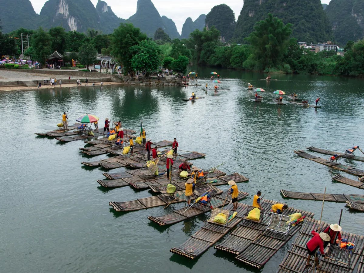 People in a calm river on rafts in China with mountains and trees on the shore in the distance. 