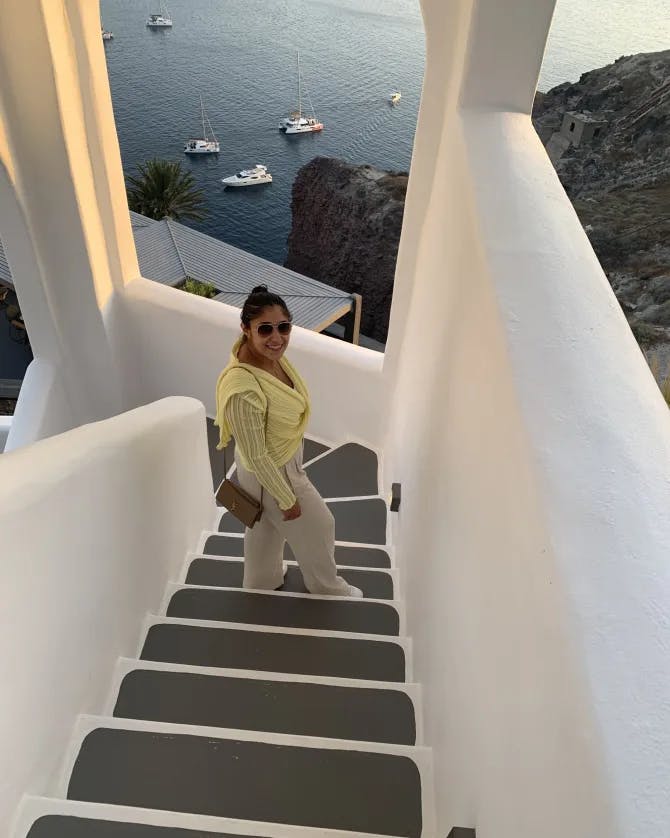 A woman in white pants, sunglasses, and a yellow top walks down a narrow stairwell overlooking the ocean.