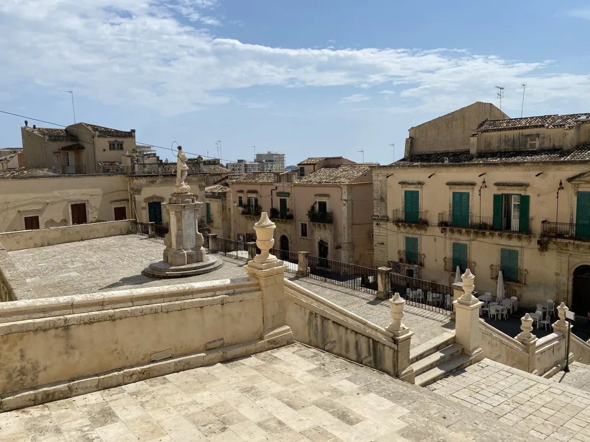 A view of Noto, Sicily showing a square with steps and a statue in the middle with surrounding old budilings.