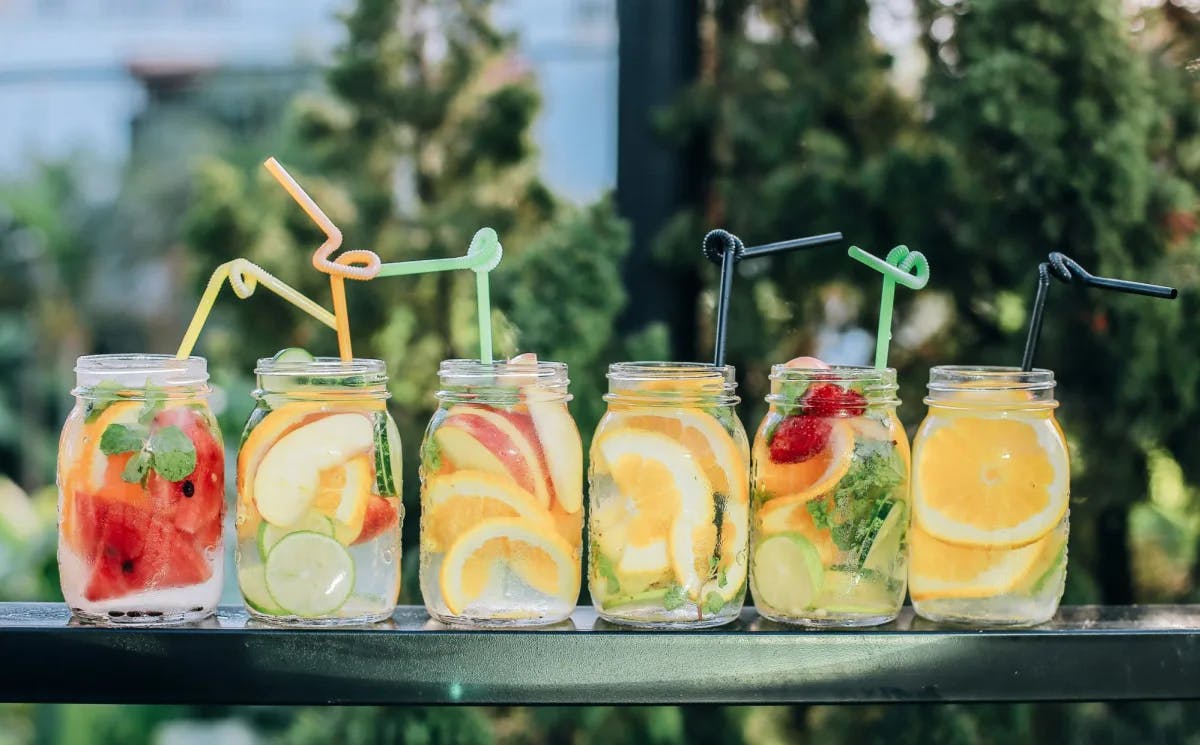 A row of cocktails with pieces of fruit in them in six mason jars and curly straws.