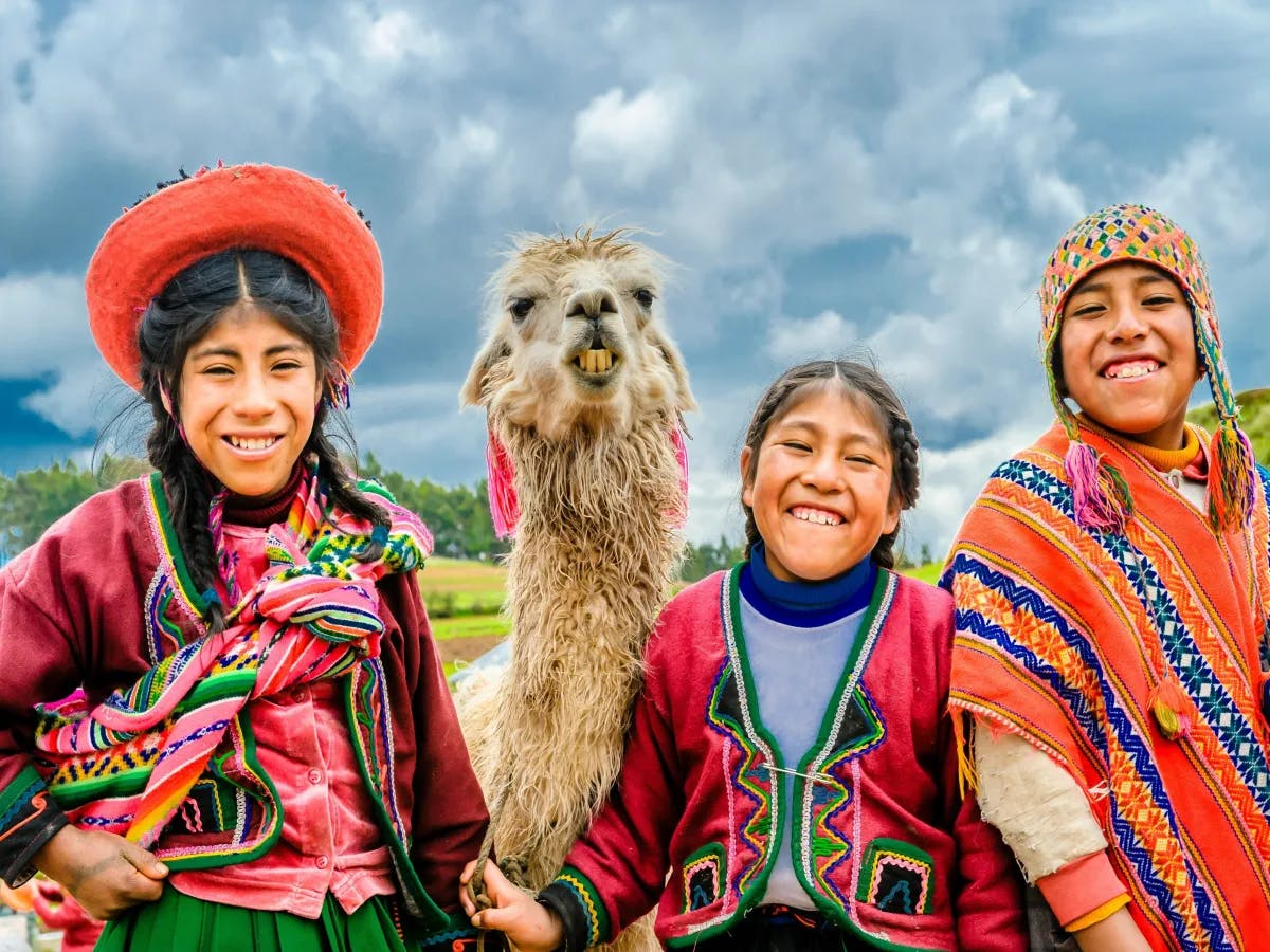 A trio in traditional Andean attire shares a moment with a llama under the vast, clouded sky.