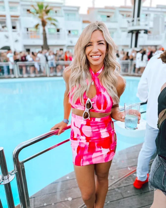 A picture of Morgan wearing a pink dress with glass in hand standing in front of a swimming pool