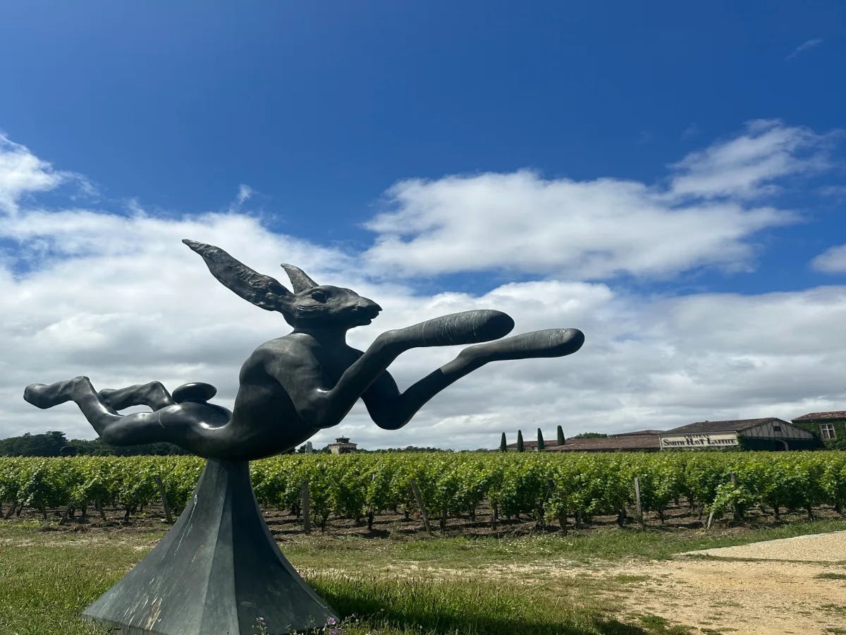 The image features a distinctive black sculpture with elongated limbs, set against a backdrop of blue sky and greenery.