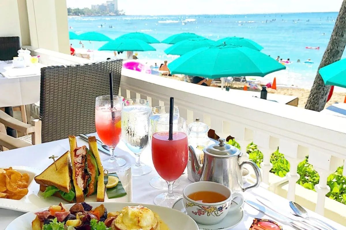 A table set up with sandwiches and drinks on a patio overlooking the beach and blue umbrellas.