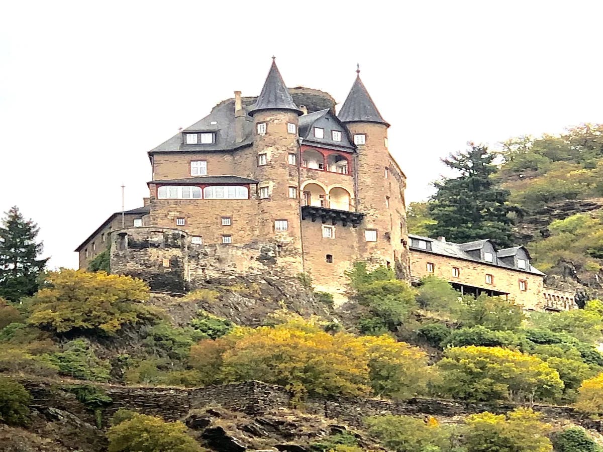 A castle perched atop a rocky cliff is surrounded by vibrant autumn foliage.