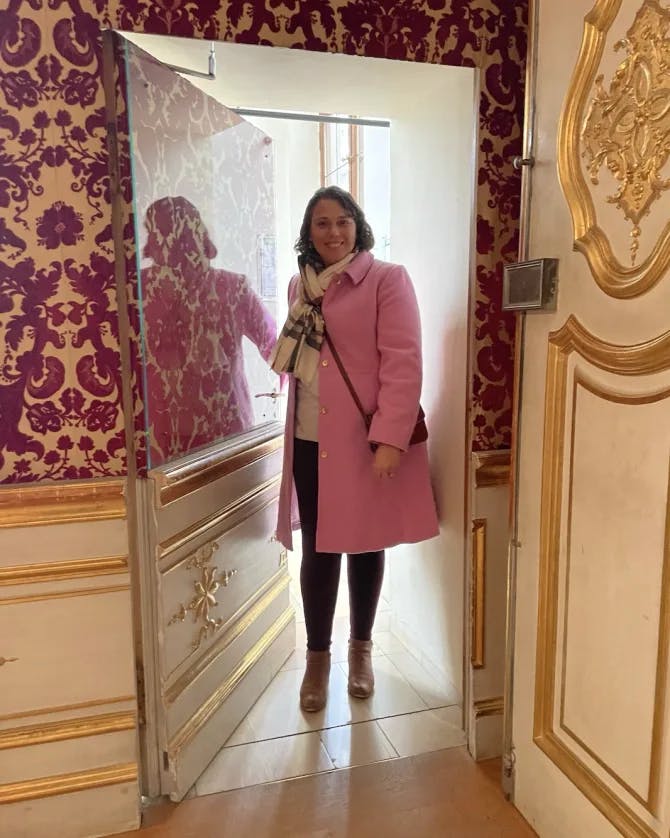 A woman in a pink blazer, white top and black pants posing in a doorway with luxurious wallpaper and gold paneled walls in the surrounding area.