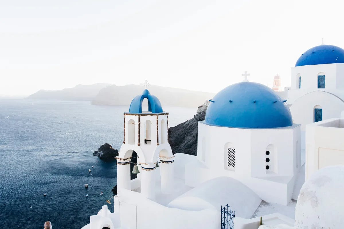 A view of the white stone buildings of Santorini with blue domed roofs. The blue sea is in the background.