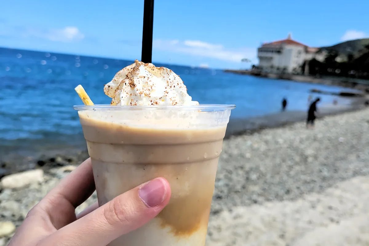 A hand holding up a frozen drink in front of a beach