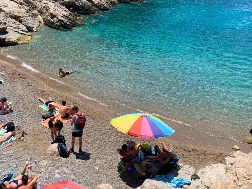 The image depicts a scenic beach setting with clear blue waters, sunbathers, and a colorful umbrella, evoking a leisurely seaside atmosphere. 