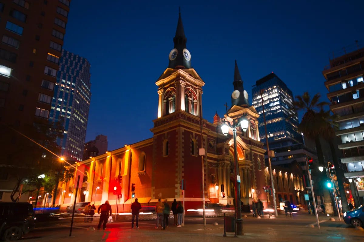 A picture of the cathedral of Santiago, Chile, illuminated at night with people walking past and other tall buildings in the background.