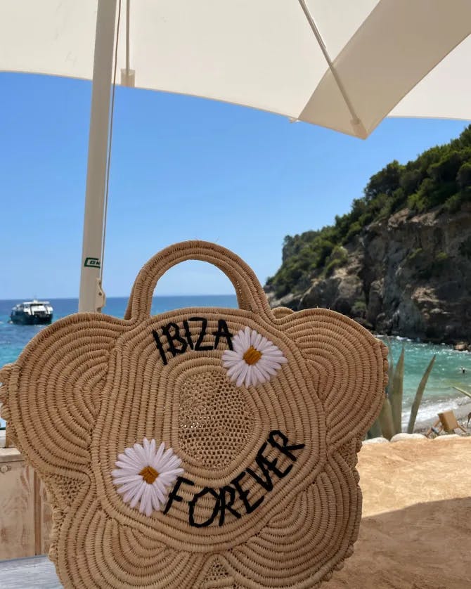 A straw purse with the embroidered text IBIZA FOREVER with a beach view behind.