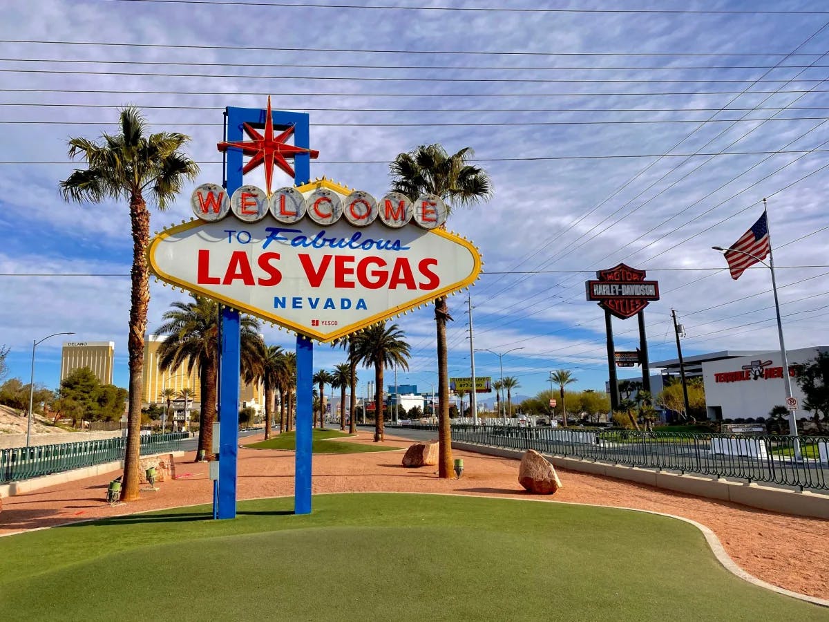 The famous sign that reads "Welcome to Las Vegas" with a green grass lawn, palm trees and an american flag.