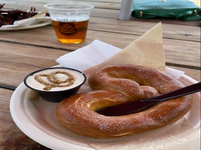 A pretzel with a side of mustard and plastic cup of beer served on a wooden table.