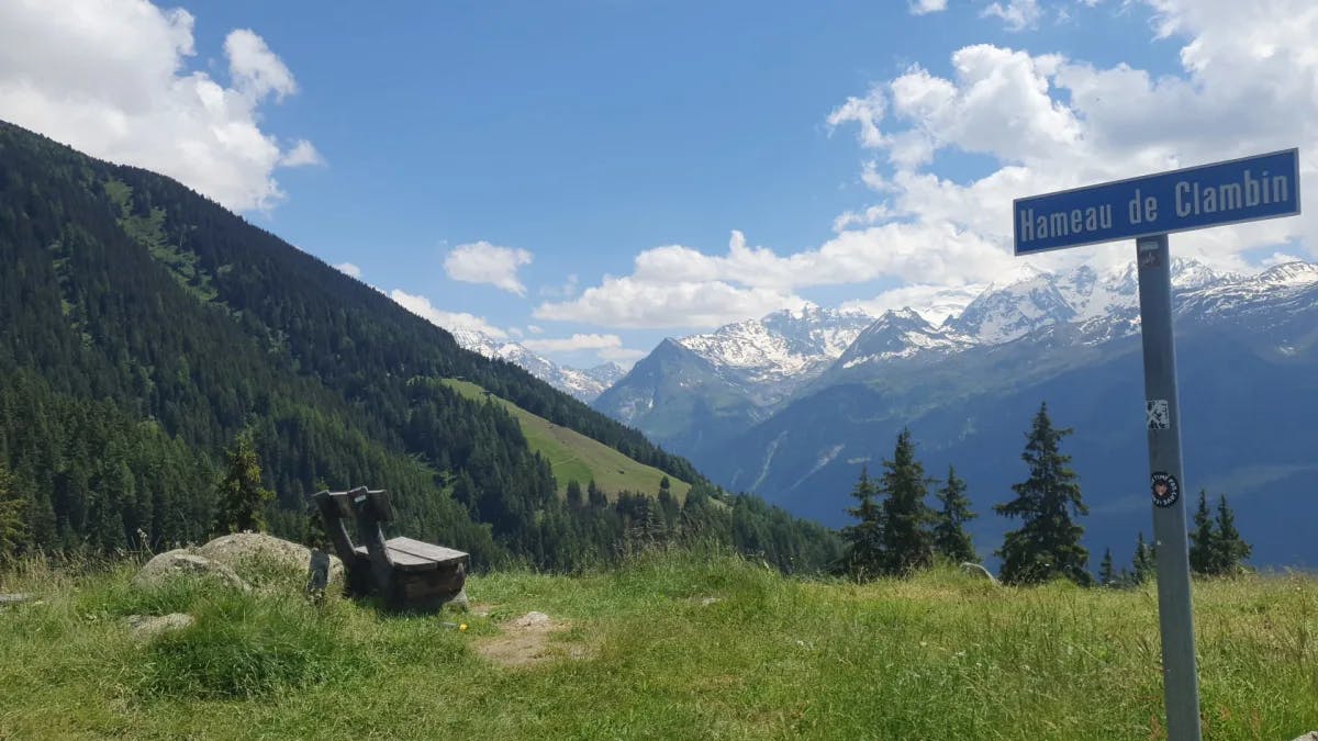 The views from a grassy clearing amidst the snow-capped mountain tops with a bench perched near an outlook spot on a clear day. 