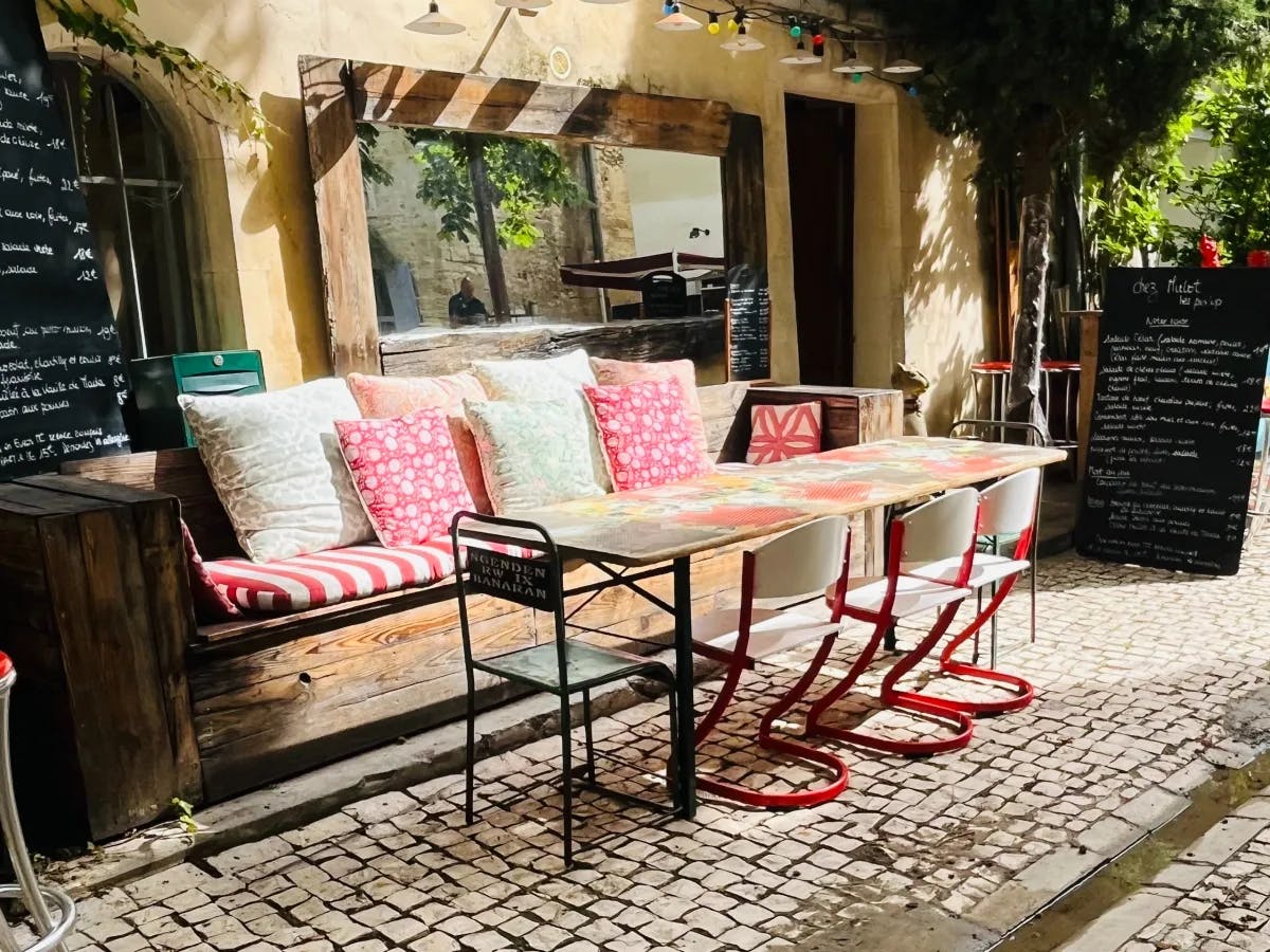 The image features a cozy outdoor cafe seating area with vibrant cushions, red chairs and a menu on blackboards.