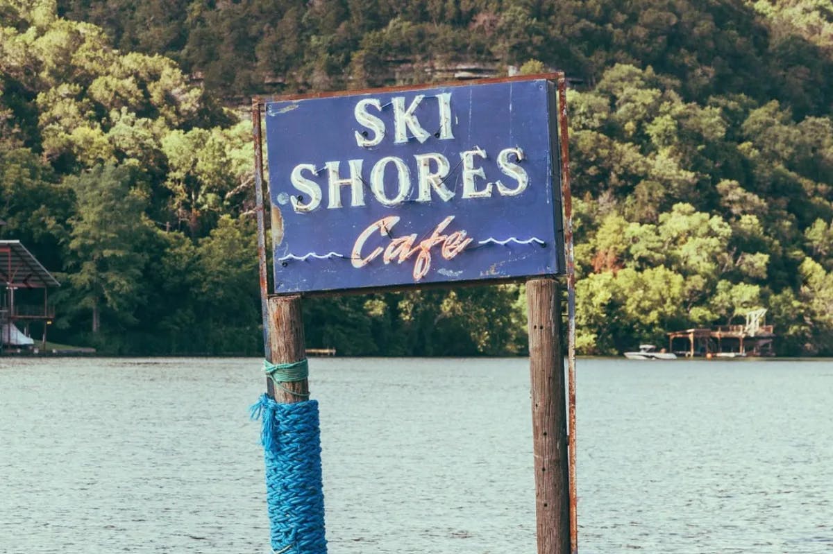 A view of a blue sign that reads "Ski Shores Cafe" over a body of water with an abundant amount of green trees in the background. 