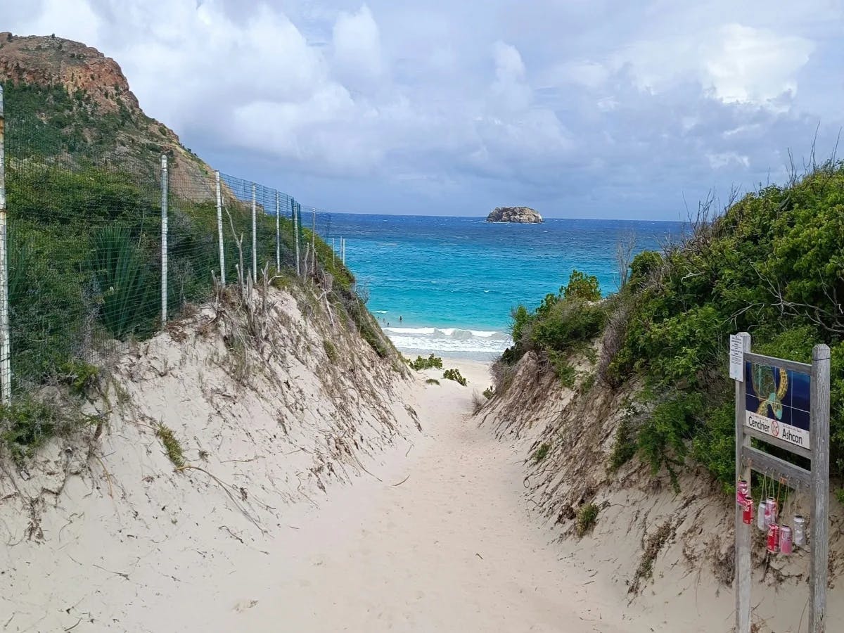 A sandy path beckons towards a beach with clear blue waters, embraced by greenery and a sky of scattered clouds.