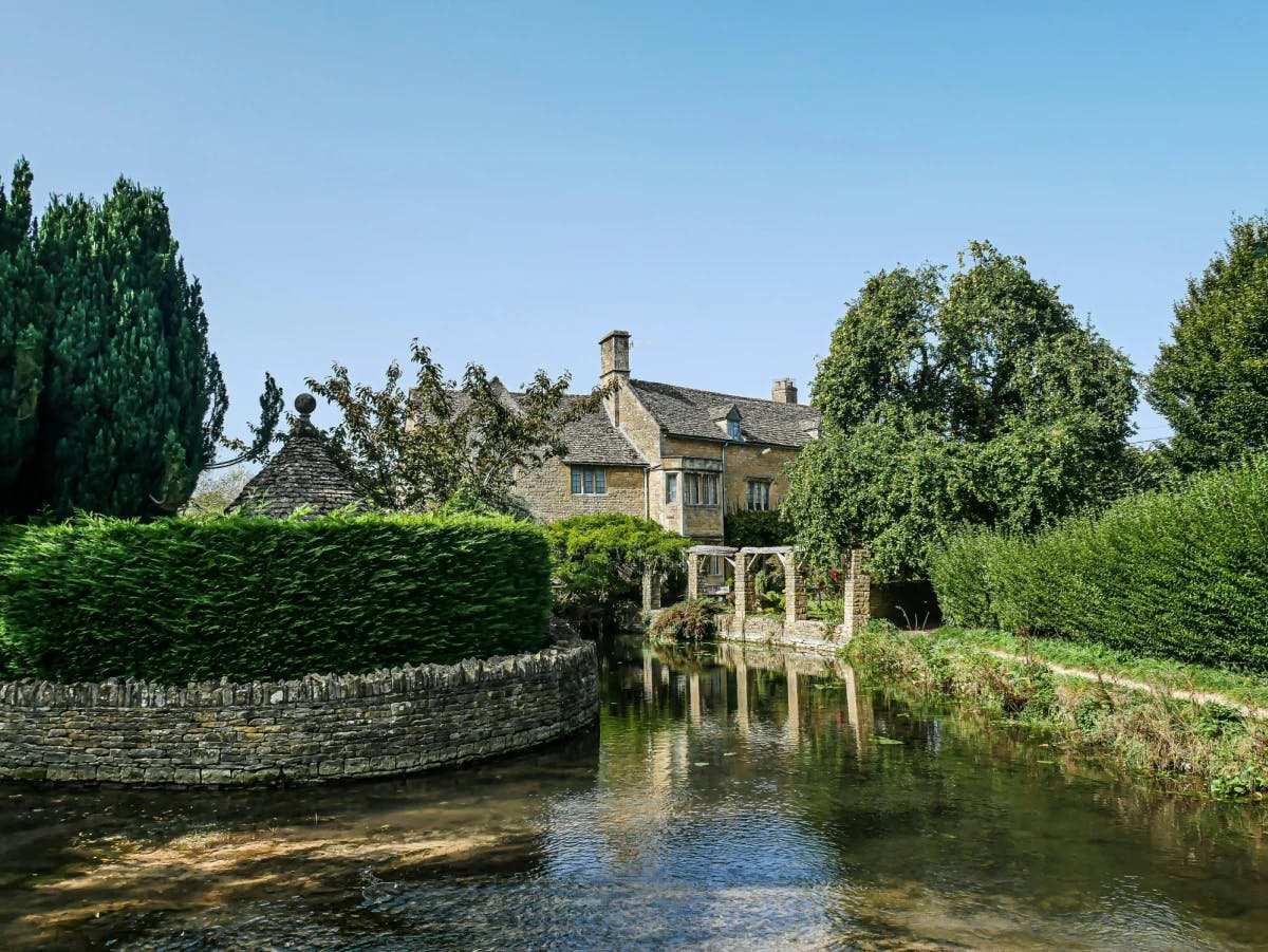 A picturesque scene of a river flowing near a stone house with bushes and greenery all around.