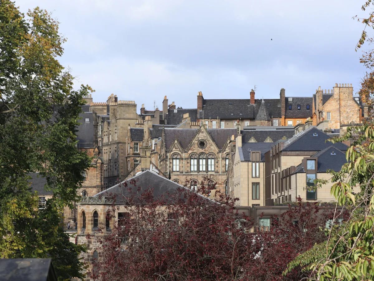 The image displays a view of historical buildings with distinctive architecture, partially obscured by trees in the foreground, under a partly cloudy sky.