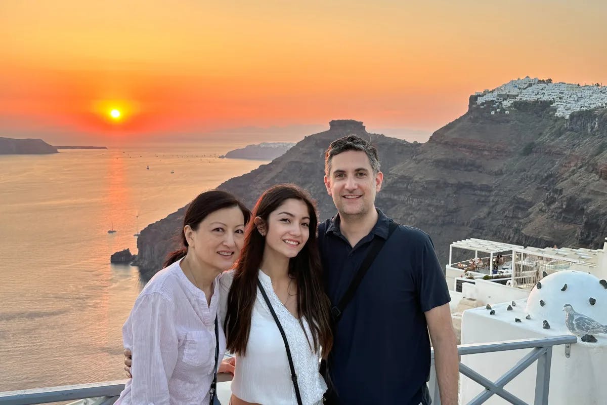 A mom, a dad, and their teenage daughter stand on a terrace overlooking an ocean sunset