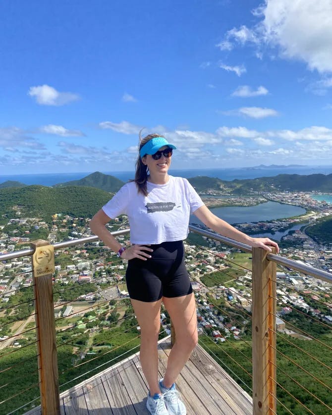 A picture of Martha wearing a white t-shirt, black shorts and blue vizor standing on an outlook that overlooks a city view and body of water