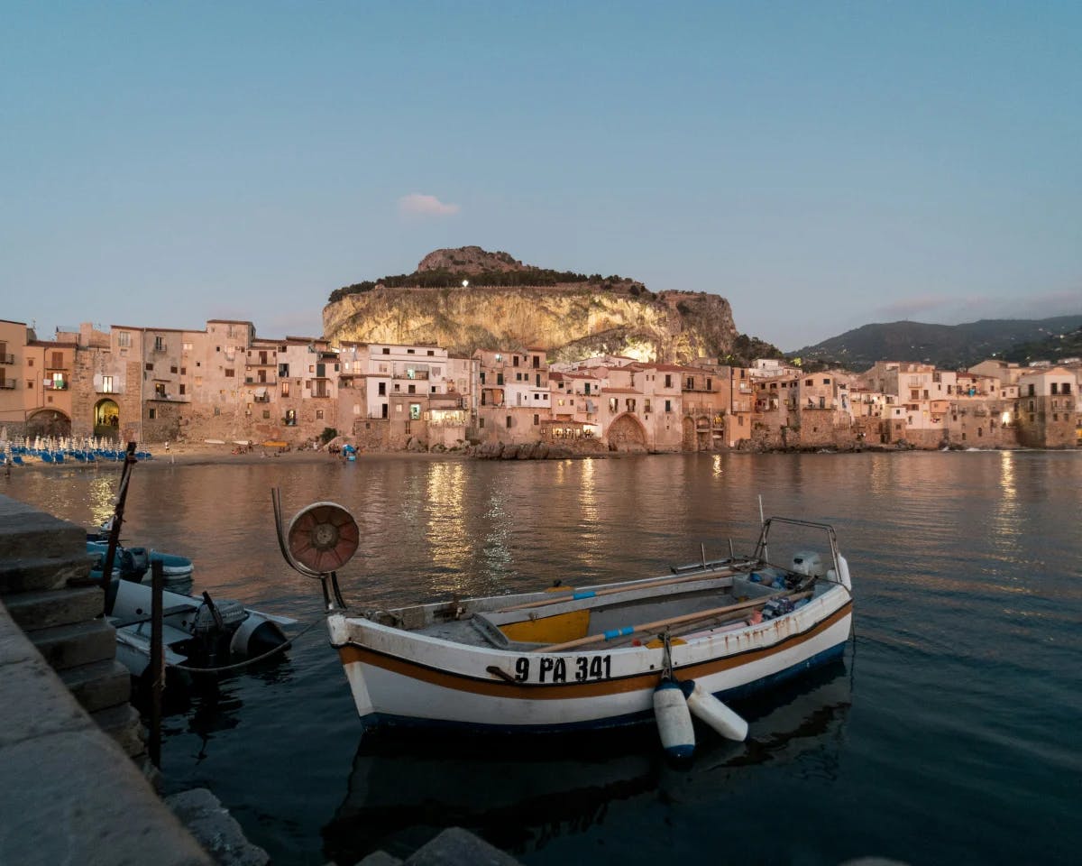 A small coastal village, with small fishing boats, buildings and houses, a mountain's peak and a small beach with people underneath a dusk sky.  