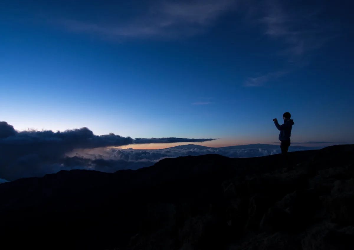 A person standing towards the right of the image outside at night with mountains, a sunset and a blue sky in the surrounding areas. 