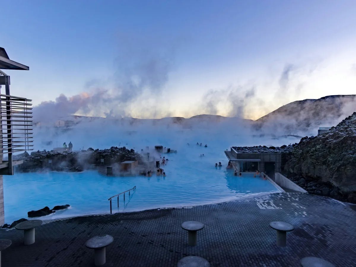 People enjoy a relaxing soak in the steaming blue waters of a geothermal spa amidst rocky terrain at dusk.