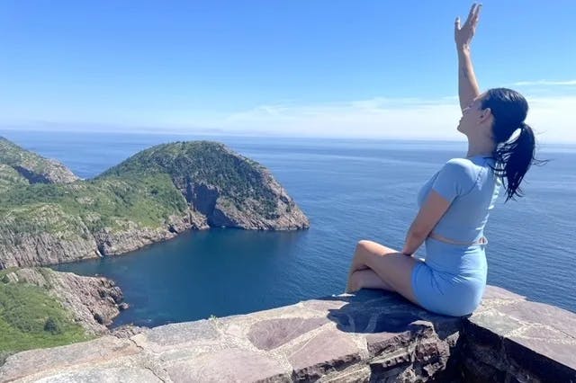 Advisor sitting on a ledge and waving while overlooking the coastline of Newfoundland.