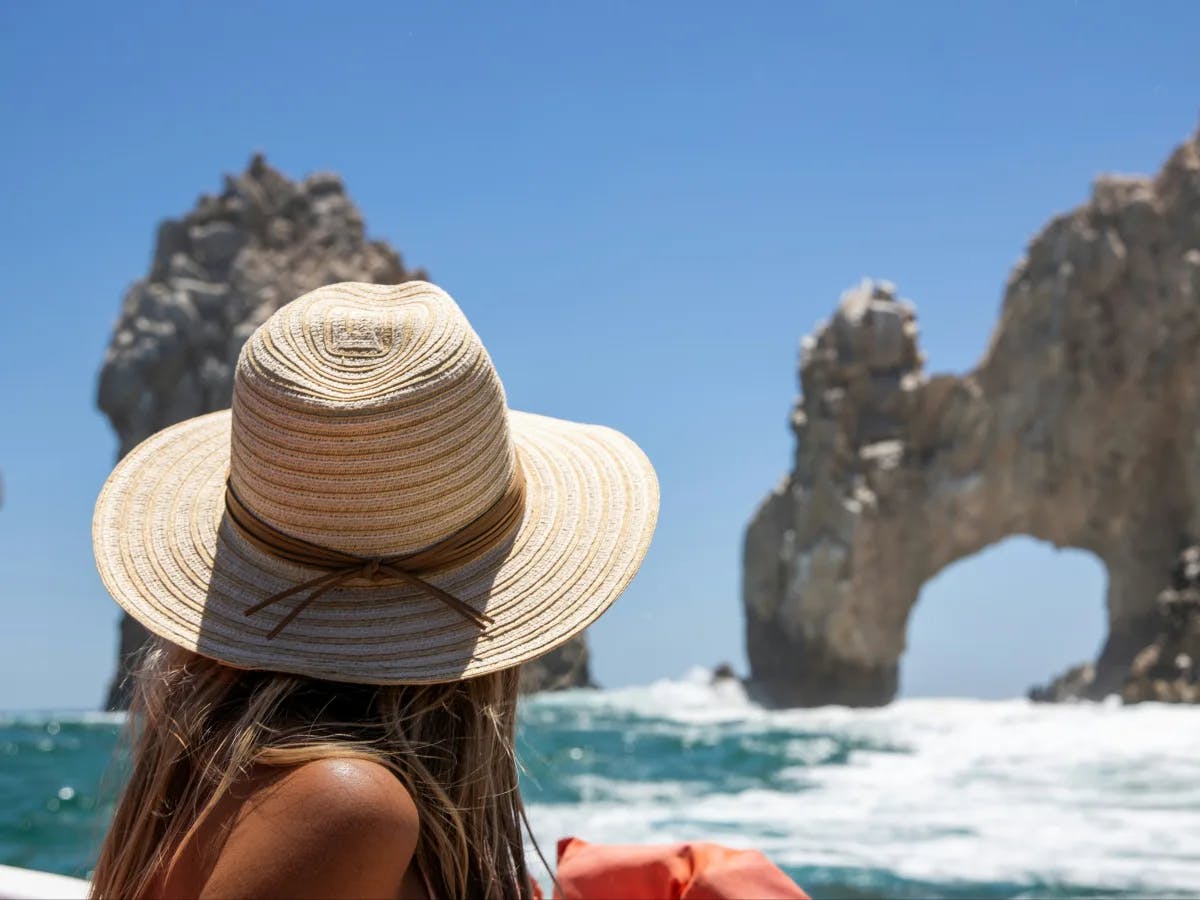 A woman in a straw hat on a boat looking at a natural rock arch in the ocean.