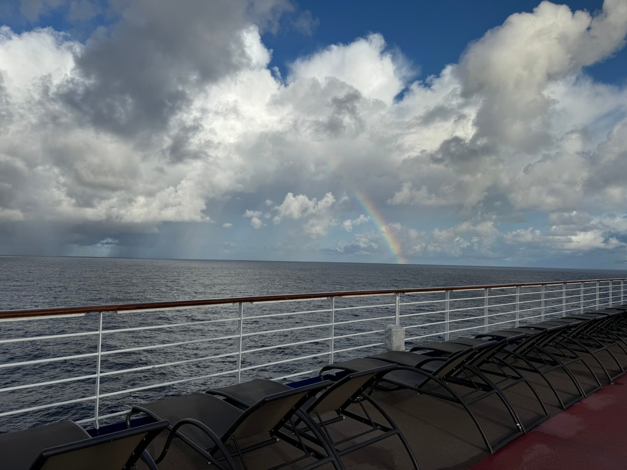 A view looking out from aboard a ship at the ocean on a partly cloudy day with a rainbow up ahead.