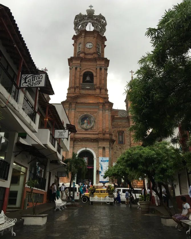 A church tower in the middle of a city square surrounded by buildings and green trees on a cloudy day 