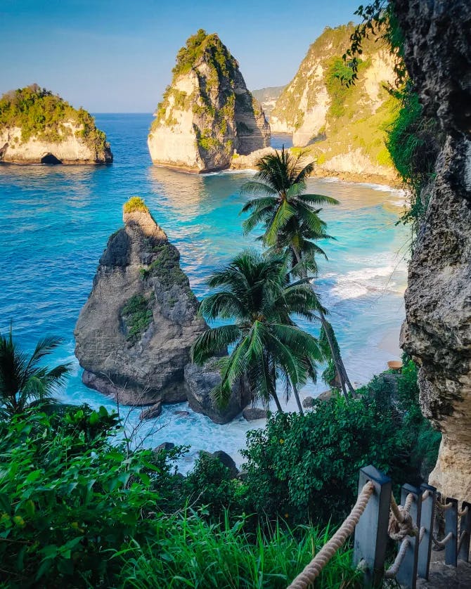 Stairs leading down to palm trees and a bright blue body of water with large rock formations spread around.